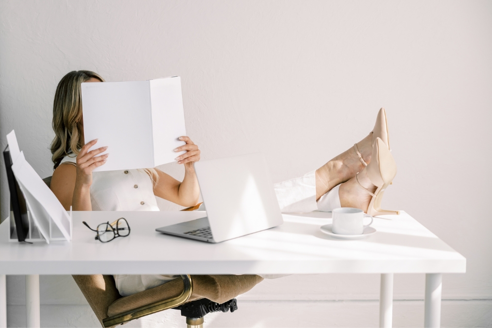 Woman reading an open book at a white desk, feet up, laptop and coffee nearby, in a bright minimal workspace